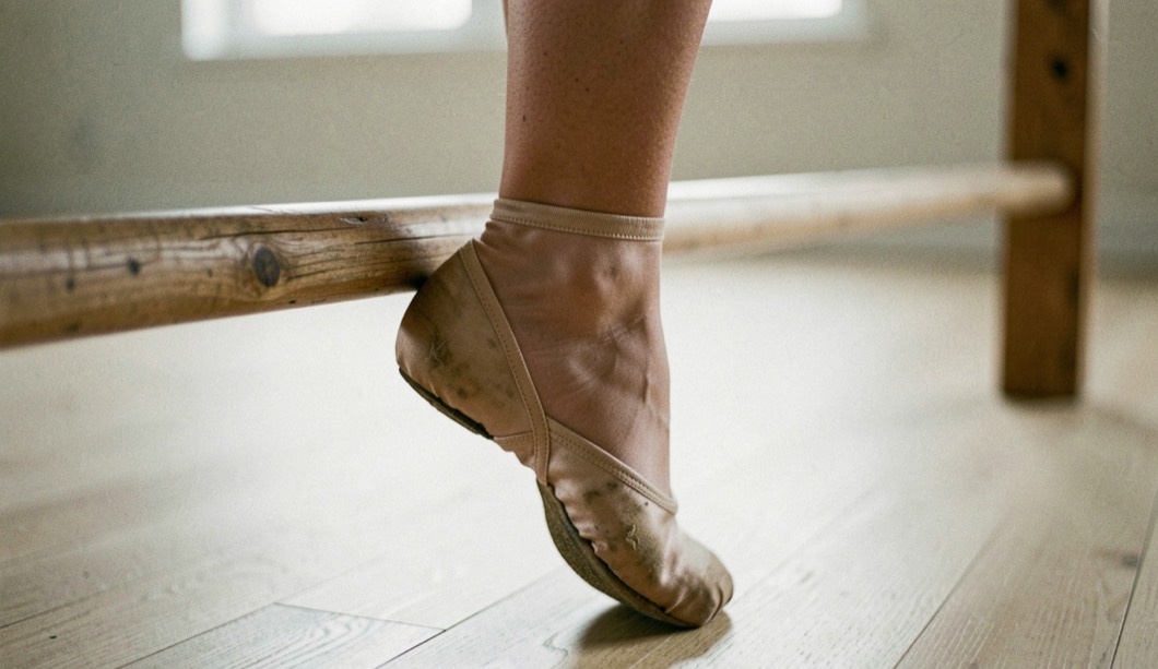 Gros plan vertical sur un cou-de-pied tendu en demi-pointe près d’une barre en bois, chausson de satin usé, parquet clair, lumière douce de studio