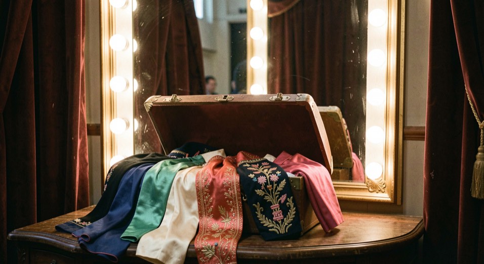 Malle de voyage ouverte dans une loge de théâtre, costumes de scène brodés et sarouels de soie débordant du couvercle, miroir à ampoules derrière, lumière chaude, cadrage vertical