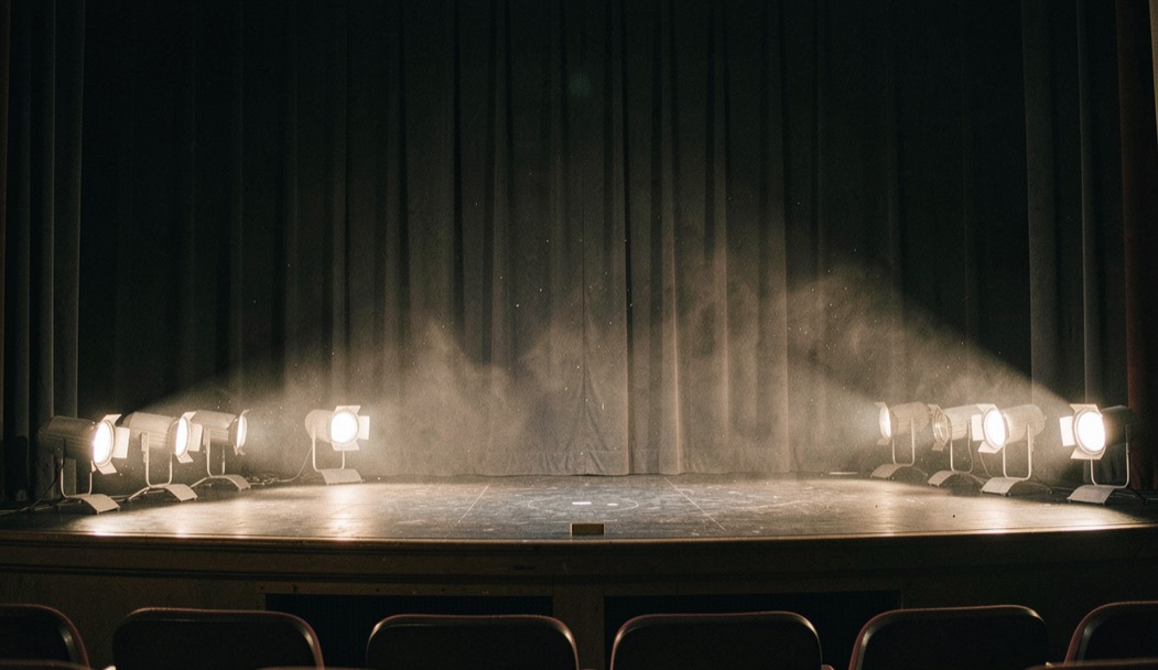 Rangée de lampes de rampe allumées au bord d’un plateau de théâtre vide, poussière en suspension dans le faisceau, rideau sombre au fond, cadrage vertical rasant pris depuis la fosse